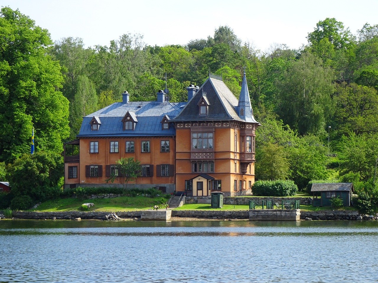 Historic orange house with blue roof by a lake surrounded by lush green trees and clear water in a serene environment
