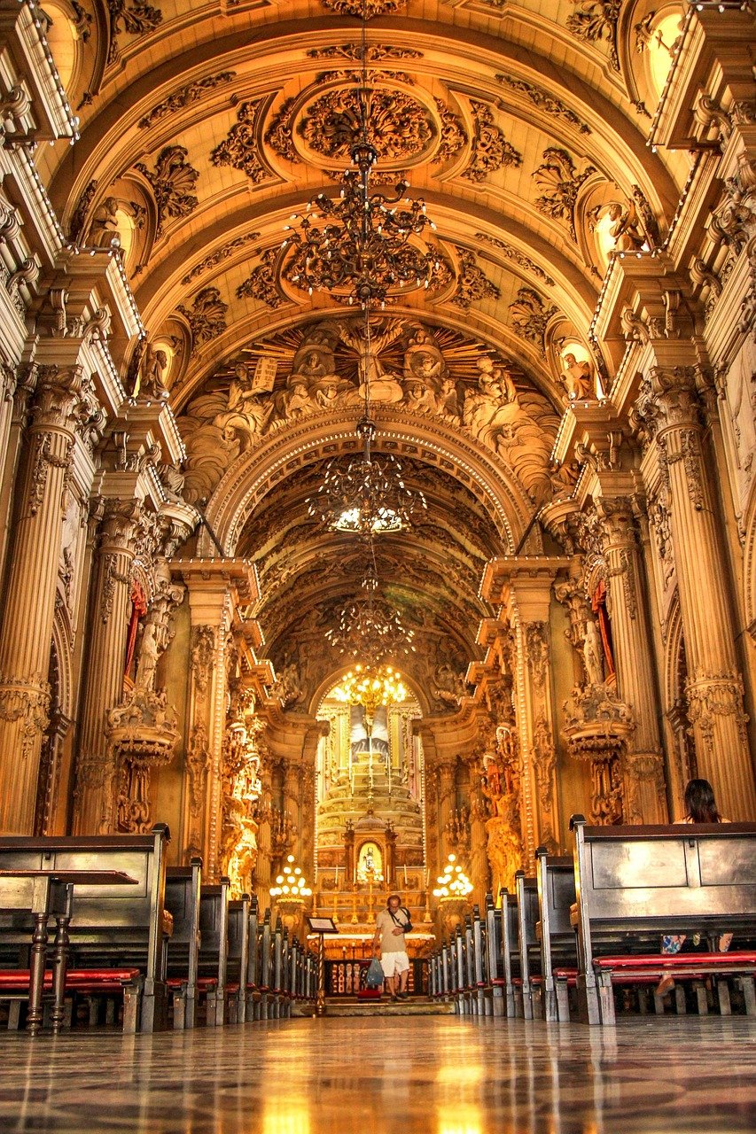Interior view of a historic ornate church with detailed arches, chandeliers, and wooden pews with visitors inside
