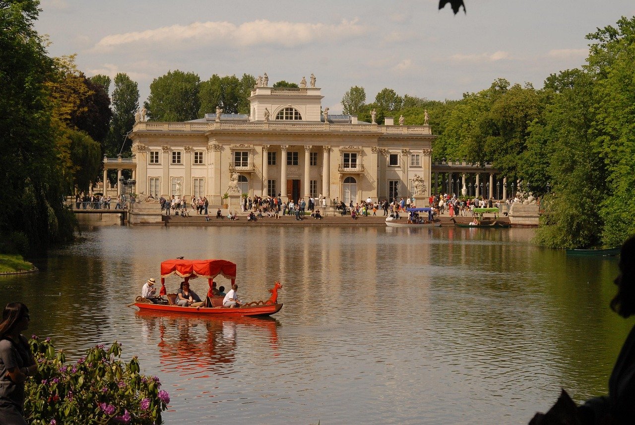 Historic palace with classical architecture by a lake with a red boat and tourists enjoying a sunny day in a green park