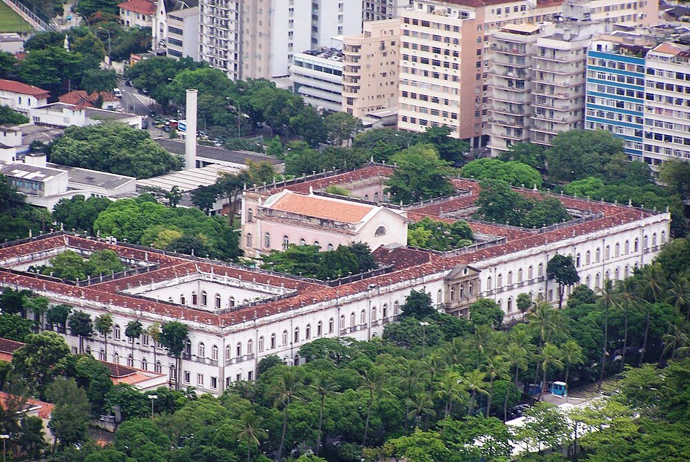 Historic white building with red roof in urban area Aerial view of a large historic white building with red roof tiles surrounded by trees and modern high-rise buildings