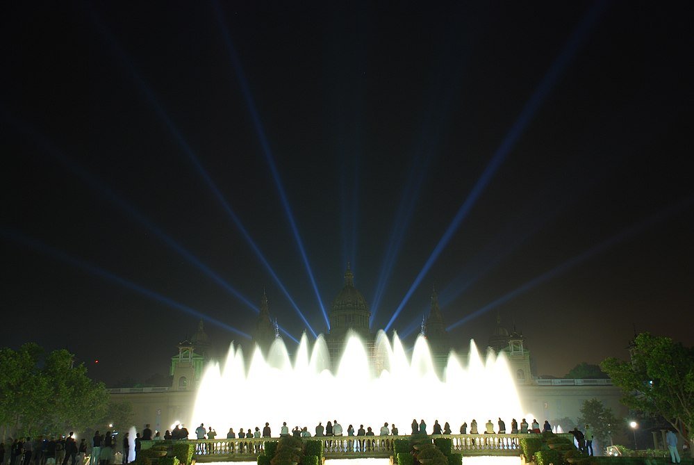 Night view of a brightly illuminated fountain with water jets and light beams in front of a historic building with people watching