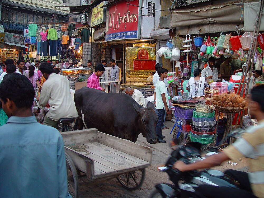 Busy Indian street market with buffalo and shops Busy Indian street market with people, a buffalo, motorbike, and various shops including a jewellers and plastic goods store