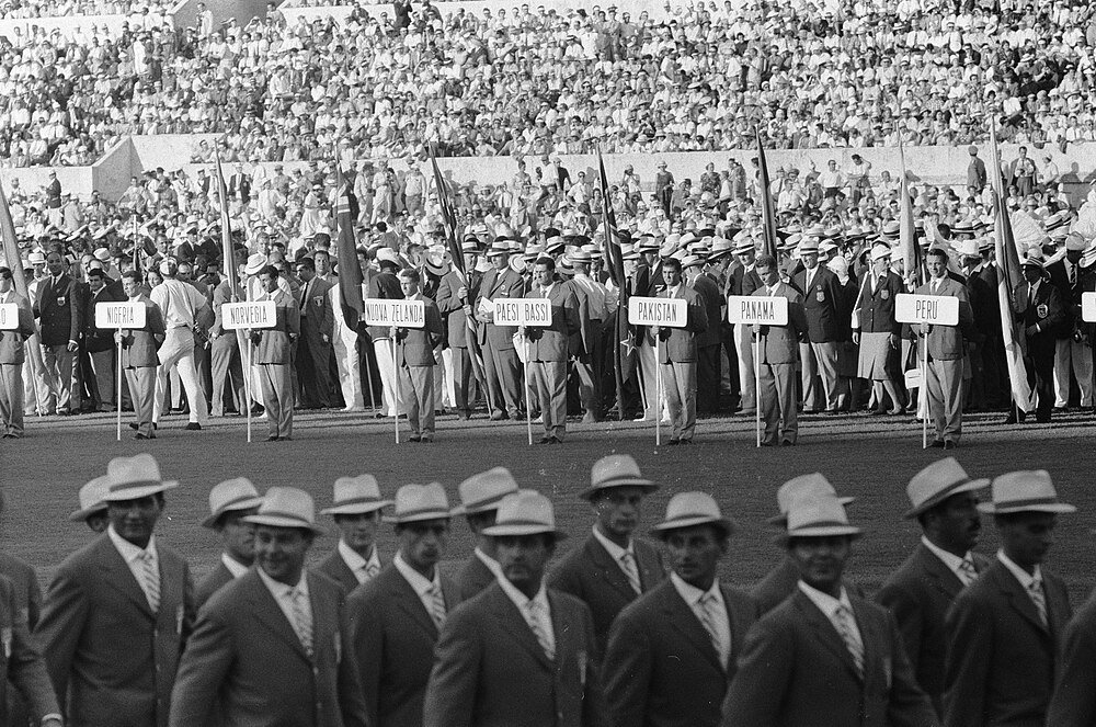 Black and white photo of international athletes marching with country signs at a large stadium event with a crowd