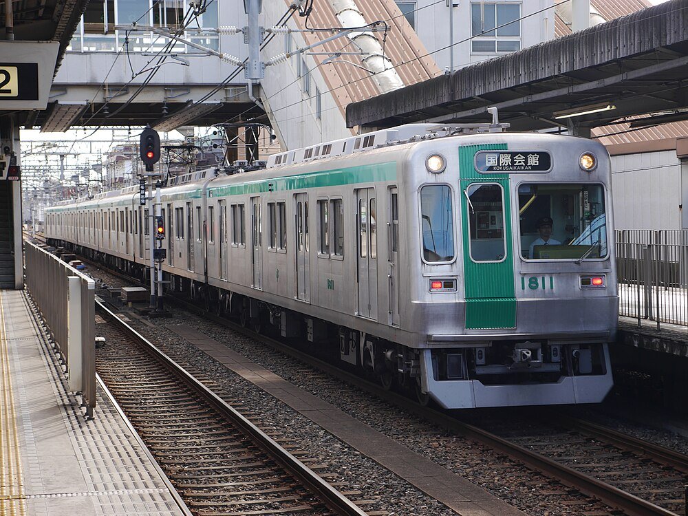 Silver and green Japanese commuter train at a station platform with overhead electric wires and signals