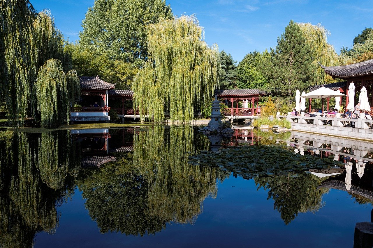 Serene Japanese garden with traditional architecture, willow trees, and calm pond reflecting the landscape on a sunny day