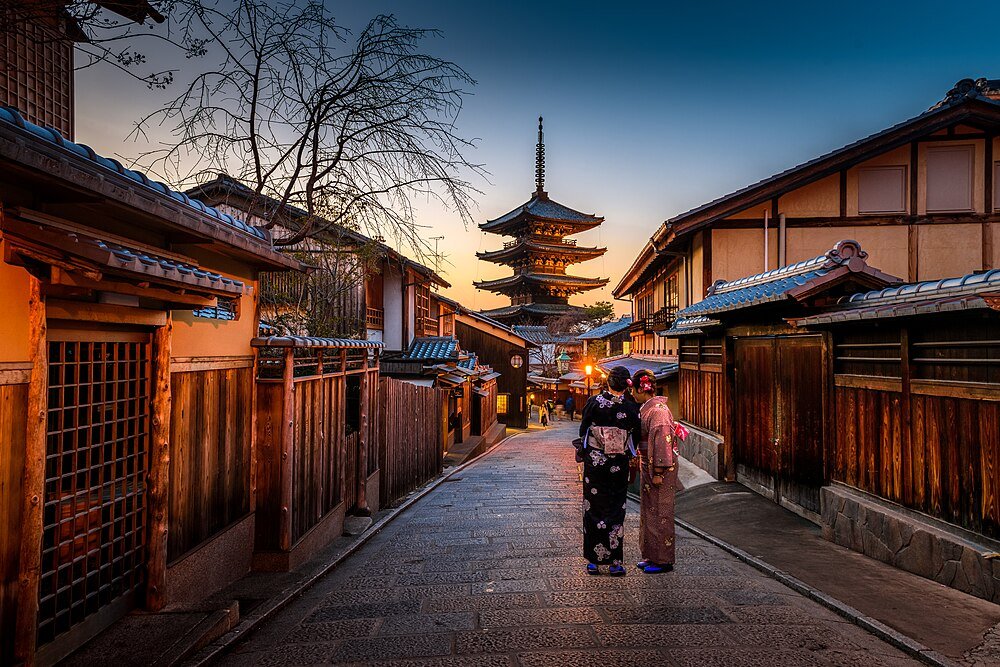 Traditional Japanese street at sunset with wooden houses, two women in kimonos, and a pagoda in the background