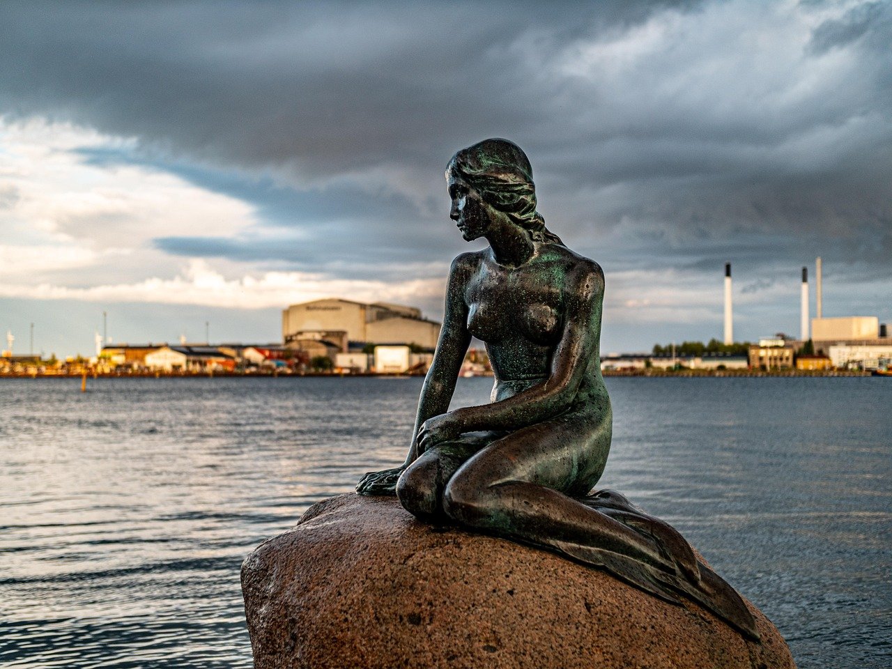 The Little Mermaid statue sitting on a rock by the water in Copenhagen with industrial buildings and cloudy sky in the background