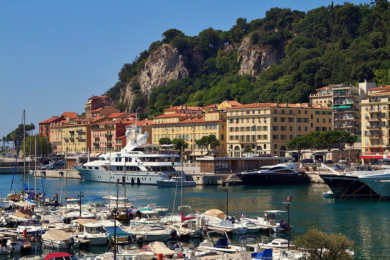 Scenic marina with luxury yachts, colorful buildings, and lush green hills under a clear blue sky