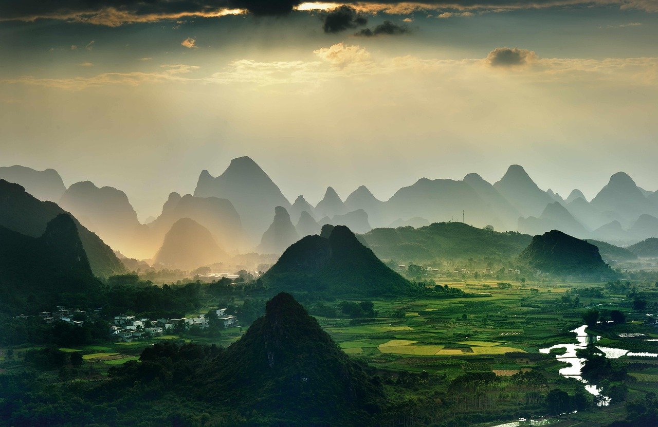 Scenic view of misty green mountains and farmland with a river flowing through a rural village at sunrise