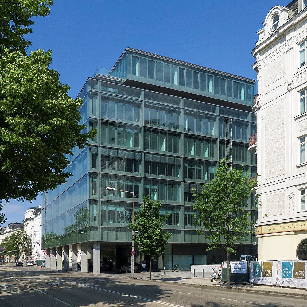 Modern glass office building with multiple floors and trees along the street under a clear blue sky