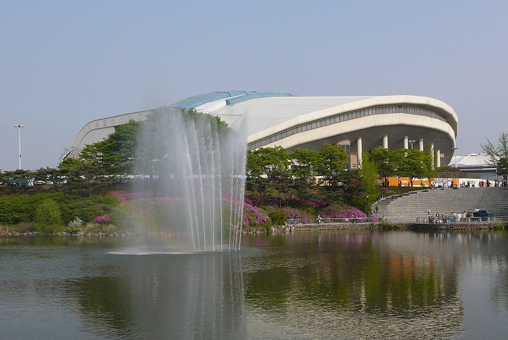 Modern stadium with water fountains and lake Large modern stadium with curved roof near a lake with water fountains and surrounding greenery and flowers