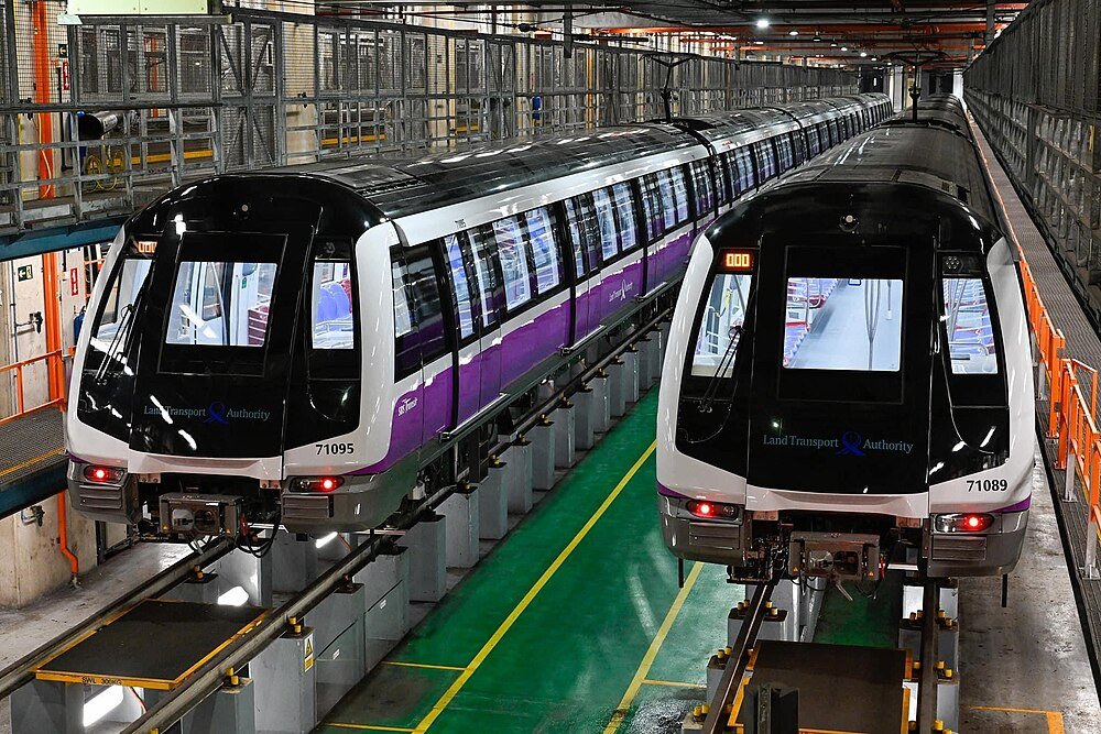 Two modern purple and white trains parked inside a maintenance depot with green flooring and metal railings