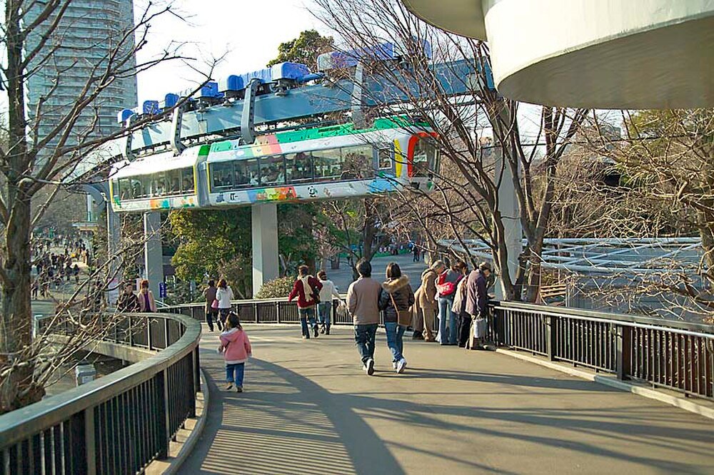 Monorail train over pedestrian walkway in urban park Monorail train passing above a pedestrian walkway with people walking and trees around in an urban park