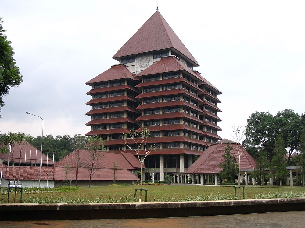 Large multi-story building with traditional red tiled roofs and pyramid-shaped top surrounded by greenery and trees