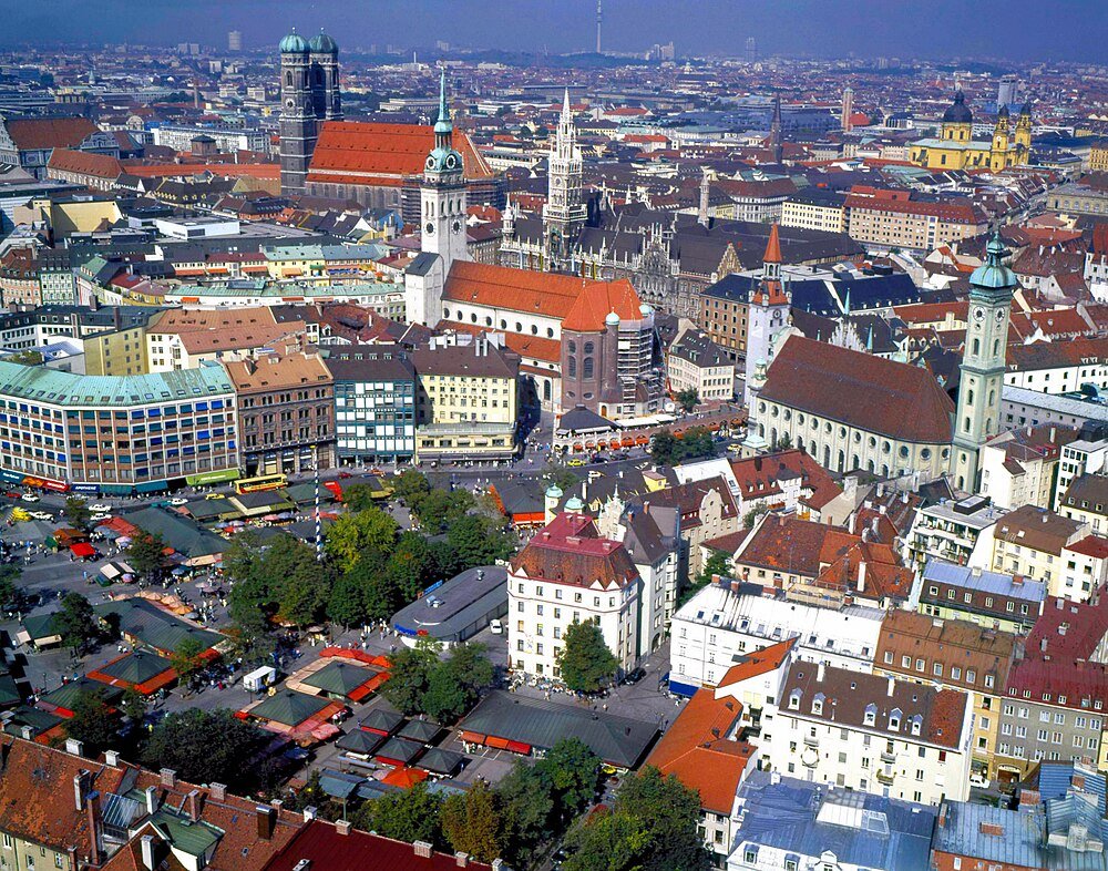 Aerial view of Munich city center with historic buildings, churches, and a bustling outdoor market under clear blue sky