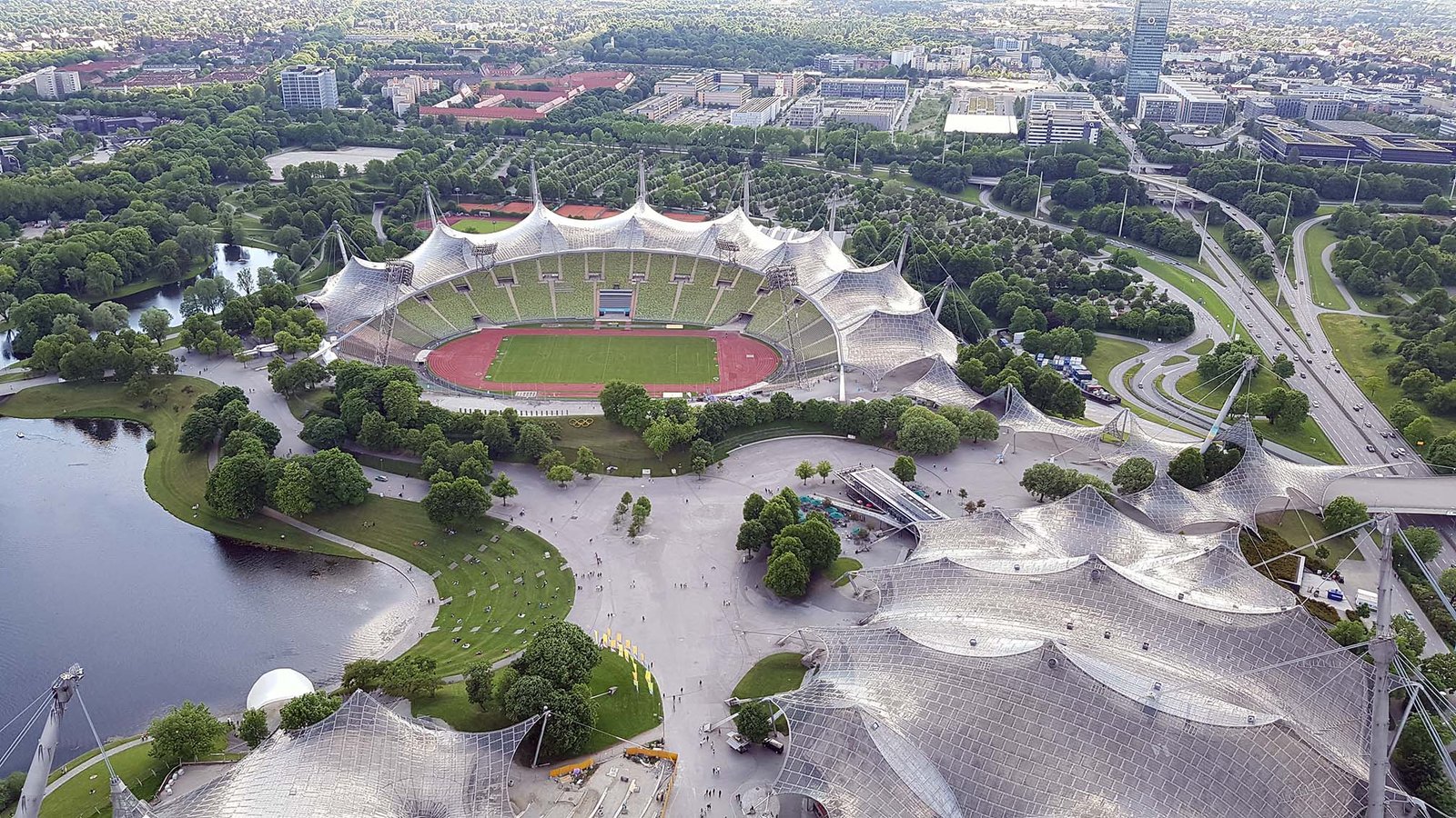 Aerial view of Munich Olympic Stadium with unique tent-like roof structure surrounded by greenery and urban roads