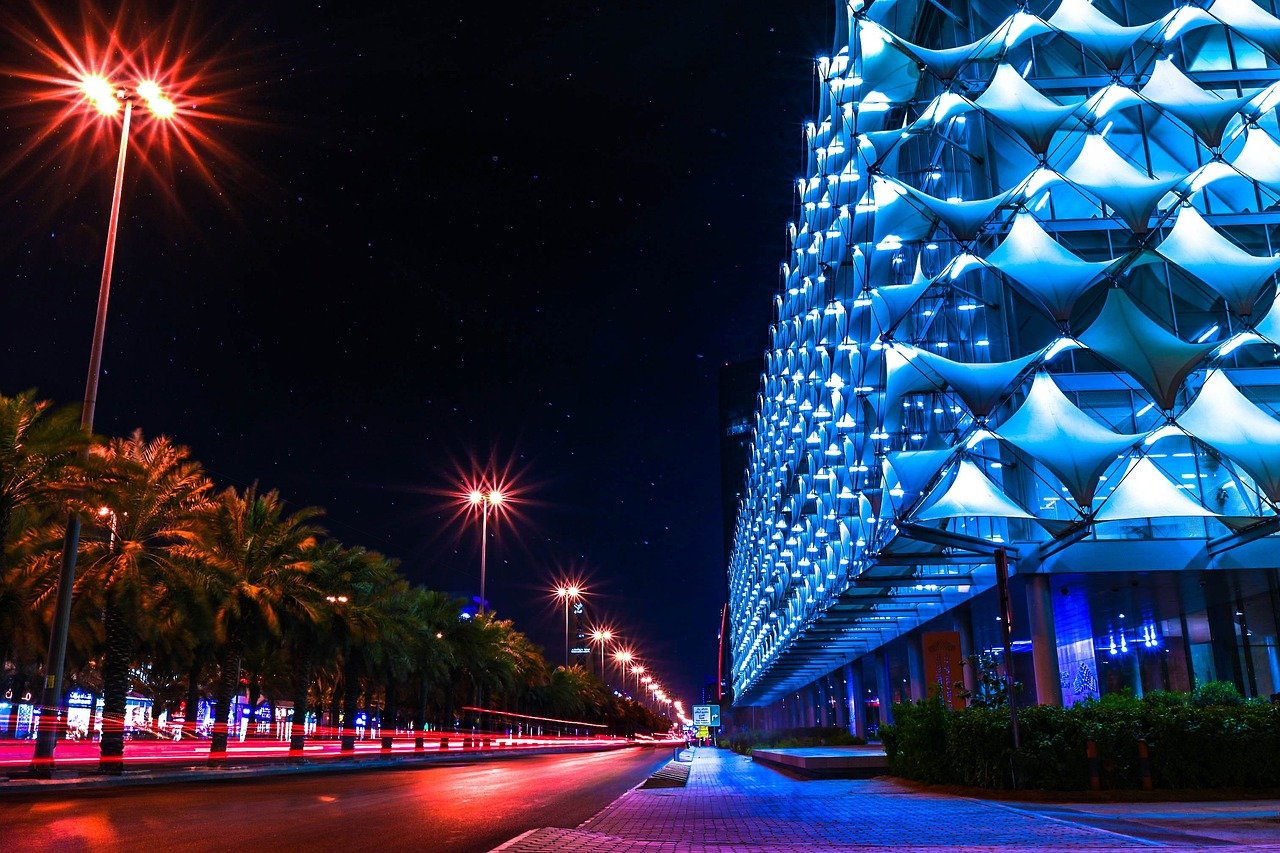 Night city street with illuminated modern building facade and palm trees under bright streetlights