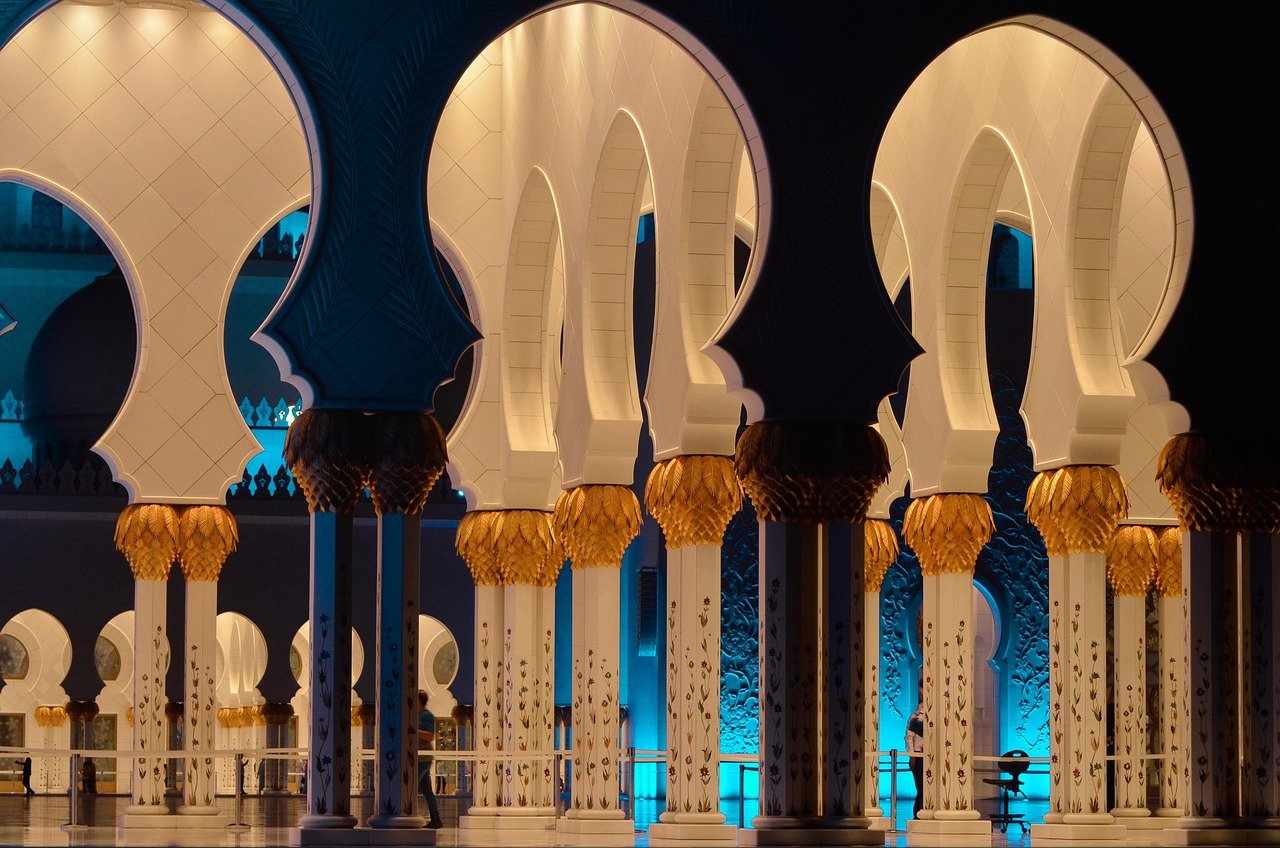 Intricately designed white and gold arches with floral patterns illuminated by warm and blue lighting in a grand mosque interior