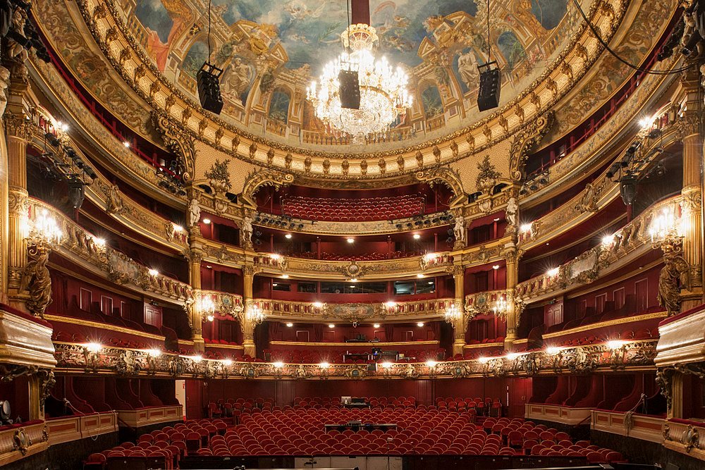 Interior view of an ornate historic theater with red velvet seats, golden decorations, and a large chandelier
