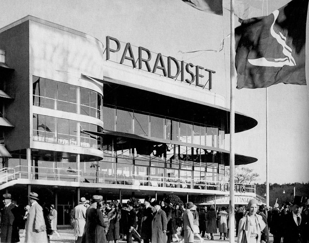 Black and white photo of a crowd outside the Paradiset building with large glass windows and flags in the foreground