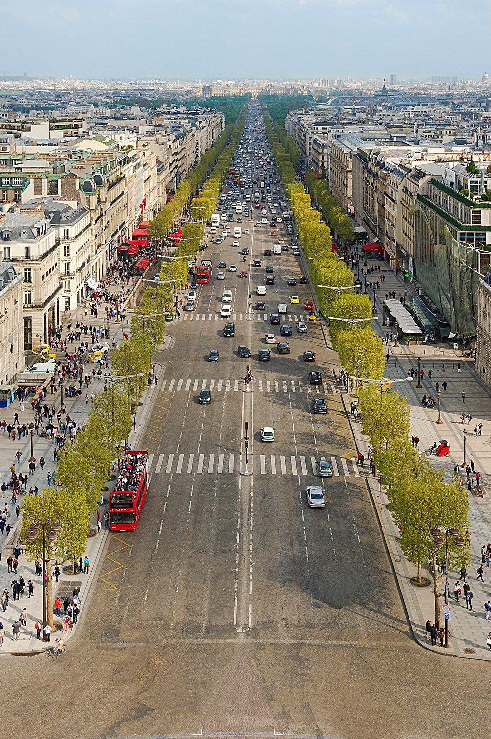 Aerial view of a busy Paris avenue lined with trees, historic buildings, and bustling pedestrian sidewalks on a sunny day