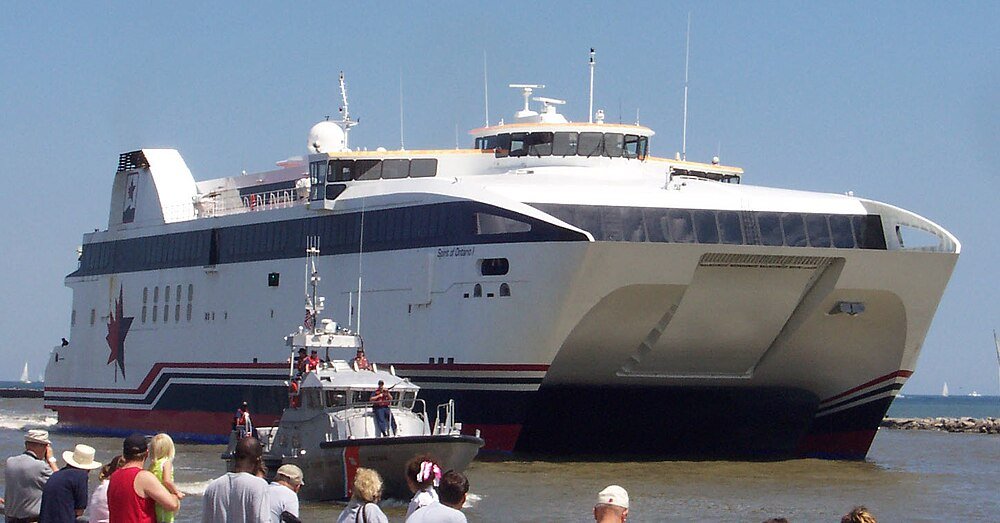 Passenger ferry boat docked near shore Large white and blue passenger ferry boat docked near shore with people watching on a sunny day