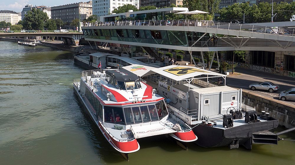 Modern passenger ferry docked alongside a floating office on a river in an urban cityscape with a bridge in the background