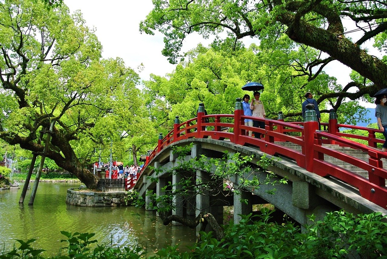 Scenic red arched bridge over calm water surrounded by lush green trees with people walking and holding umbrellas