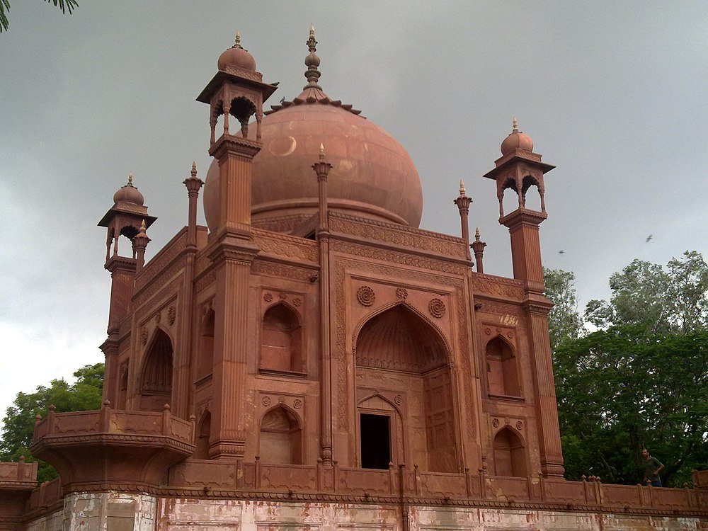 Red sandstone mausoleum with large dome and intricate carvings under cloudy sky surrounded by green trees