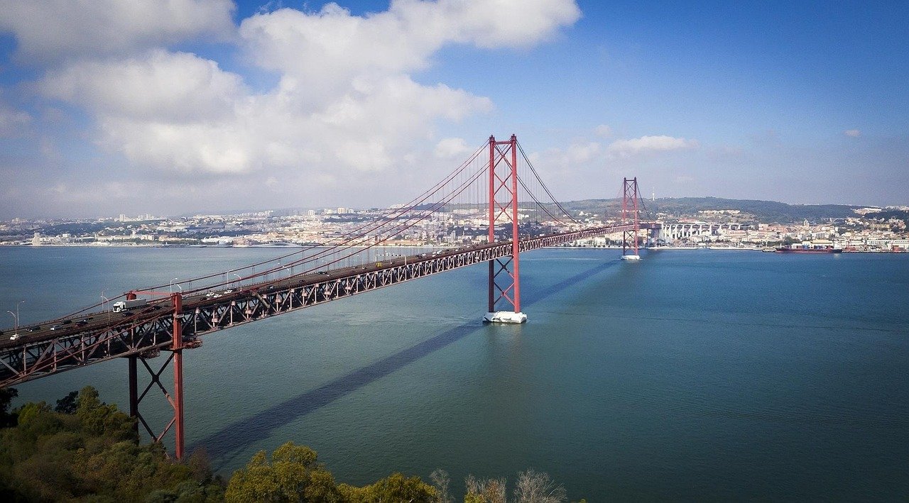 Long red suspension bridge over calm blue water with city skyline and hills in the background under a partly cloudy sky