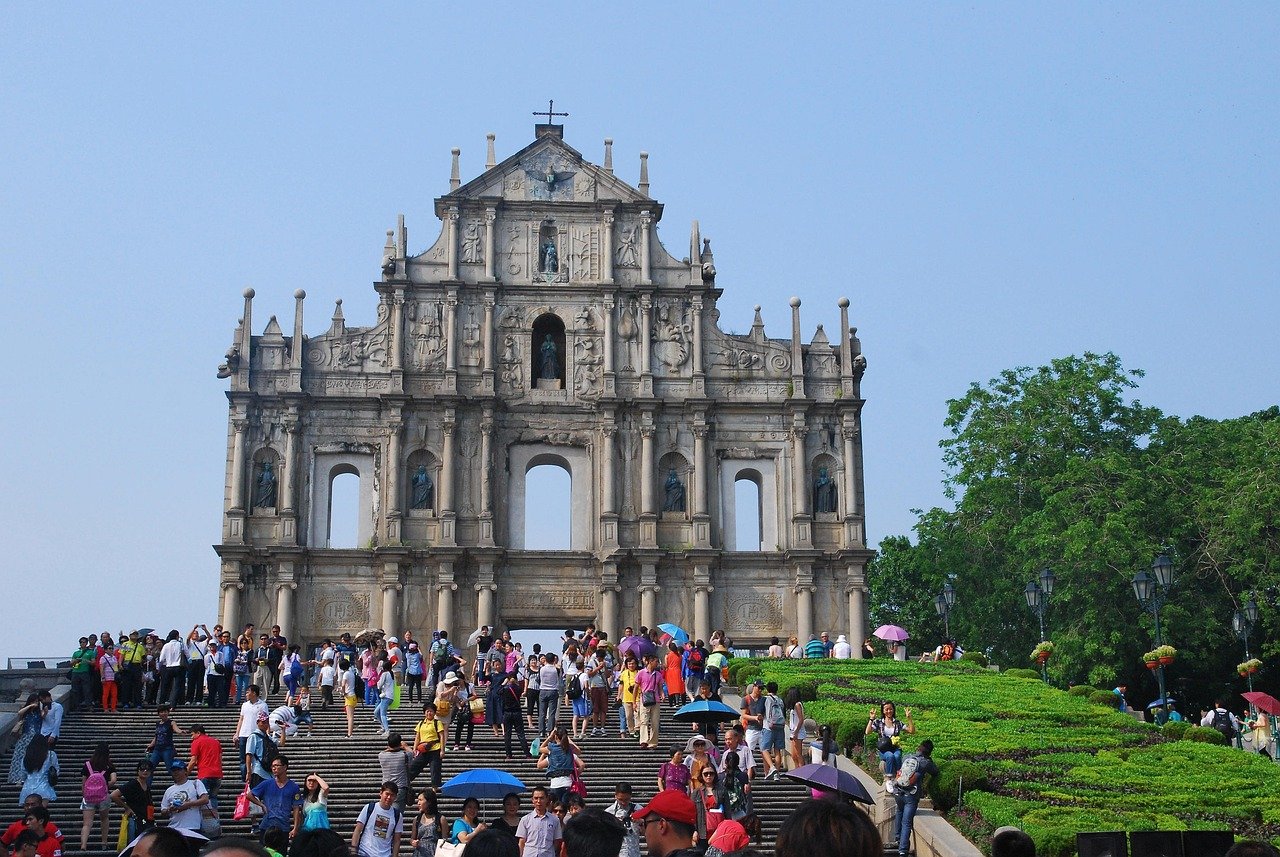 Ruins of St. Paul's historic stone facade with tourists and greenery under clear blue sky in Macau