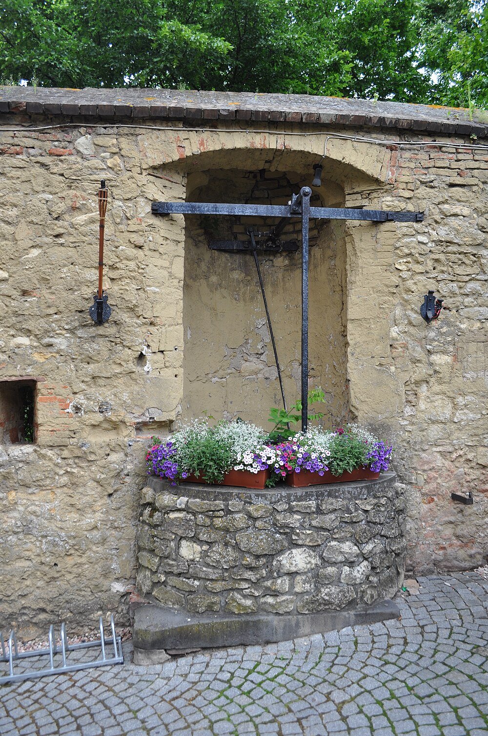 Old stone well with colorful flowers in pots on top, set against a rustic brick and plaster wall with greenery above
