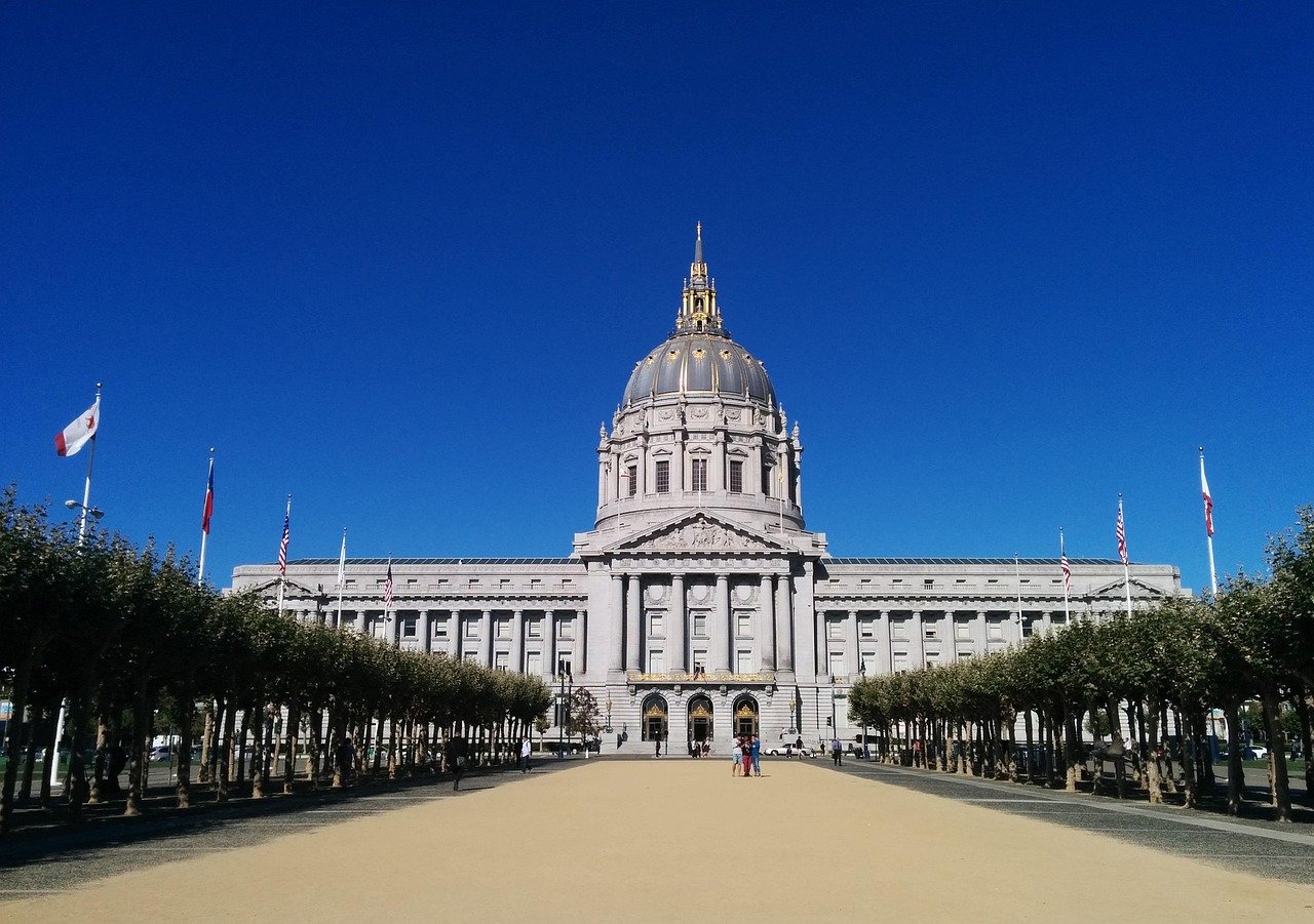 San Francisco City Hall with grand dome and flags under clear blue sky, surrounded by trees and open plaza