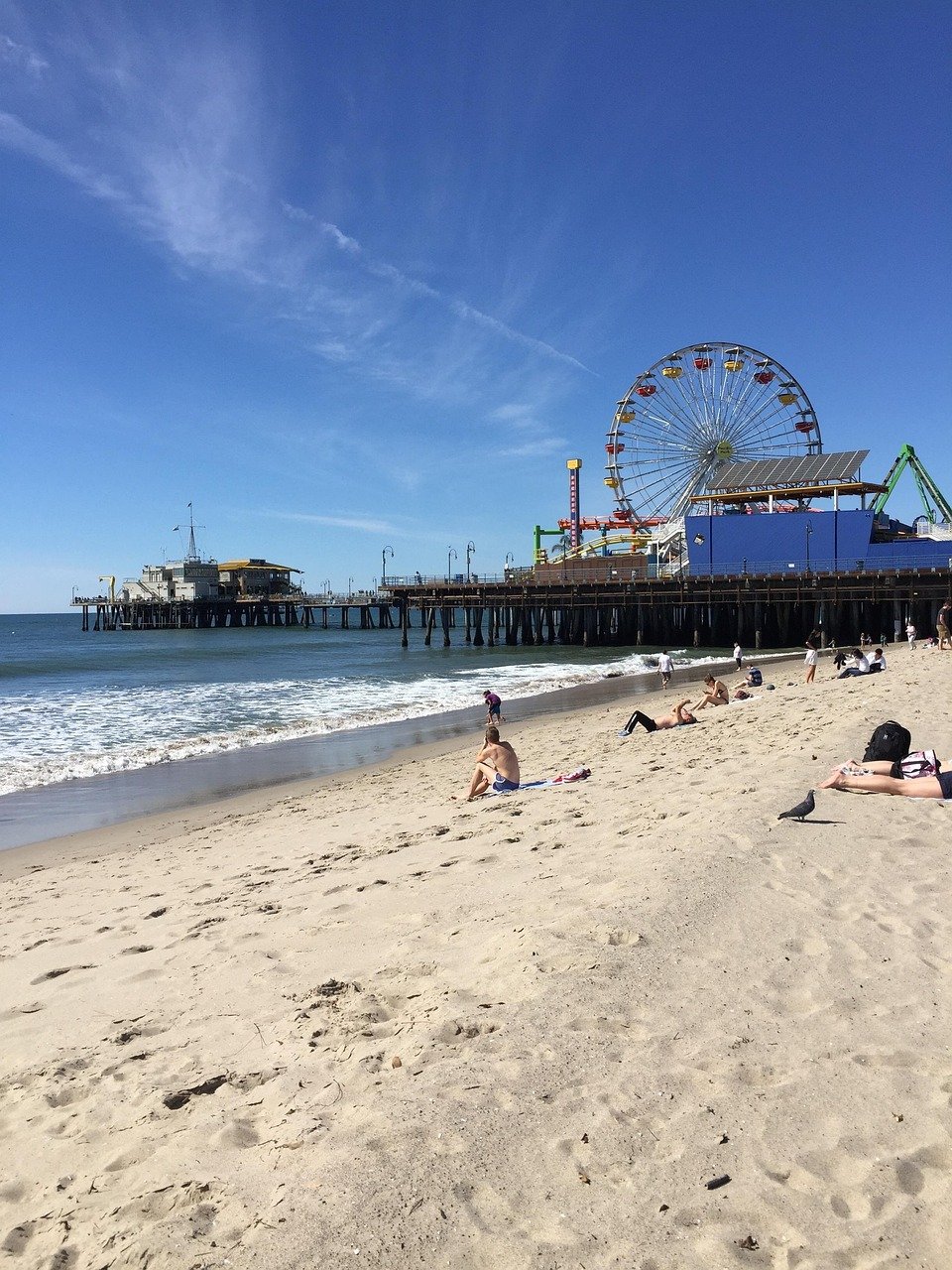 Sunny beach with people relaxing on sand near ocean and iconic Santa Monica Pier with Ferris wheel under clear blue sky