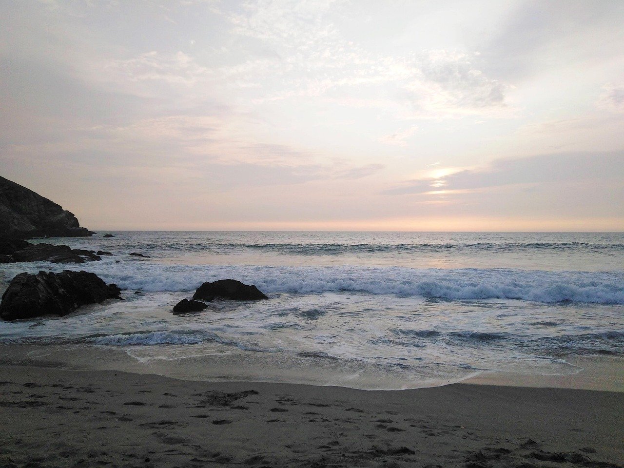 Calm ocean waves gently crashing on a sandy beach with rocks during a serene sunset under a cloudy sky
