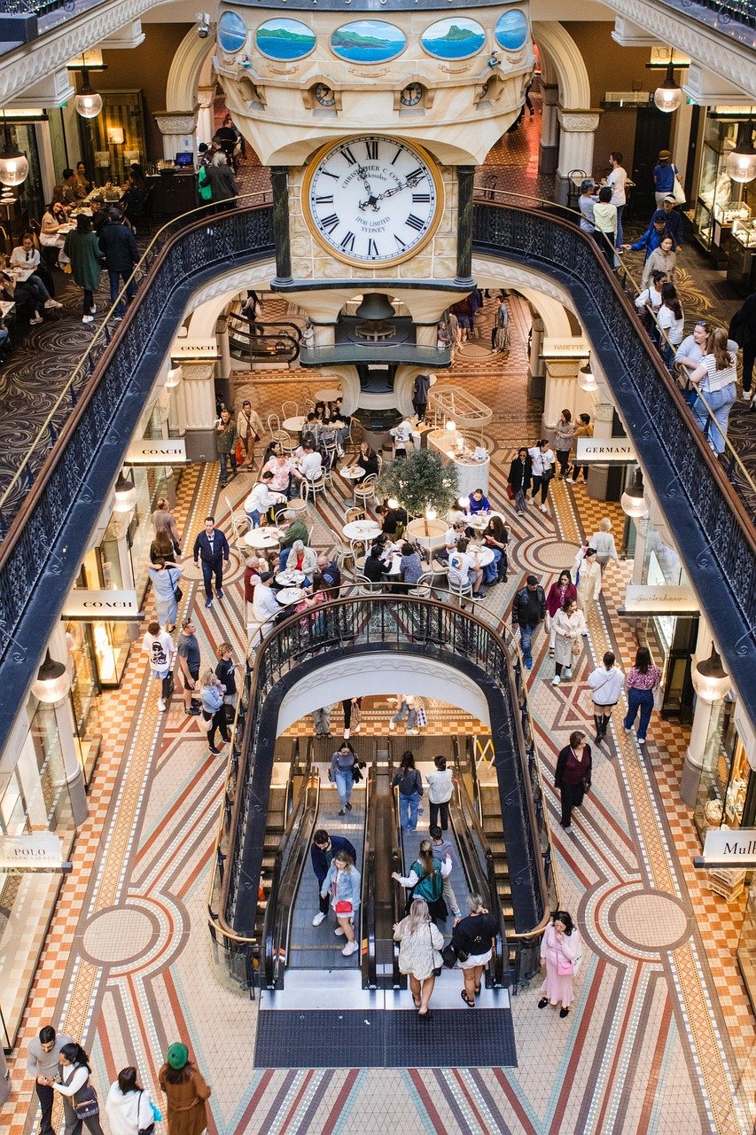 Interior view of a busy shopping mall with a large clock, escalators, and people walking and dining