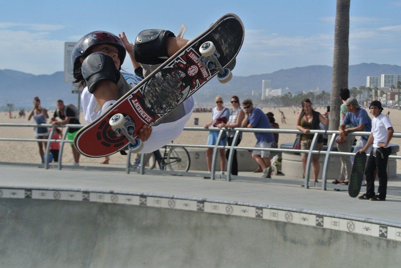Skateboarder performing an aerial trick at a sunny beach skate park with spectators and mountains in the background