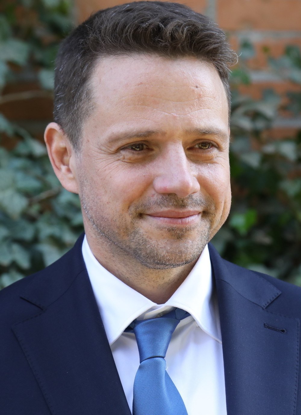 Smiling man in a navy suit and blue tie outdoors with green foliage background, professional headshot