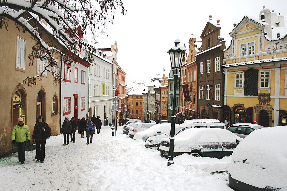 Snowy European street with historic buildings Snow-covered street with parked cars and people walking near colorful historic buildings in a European town during winter