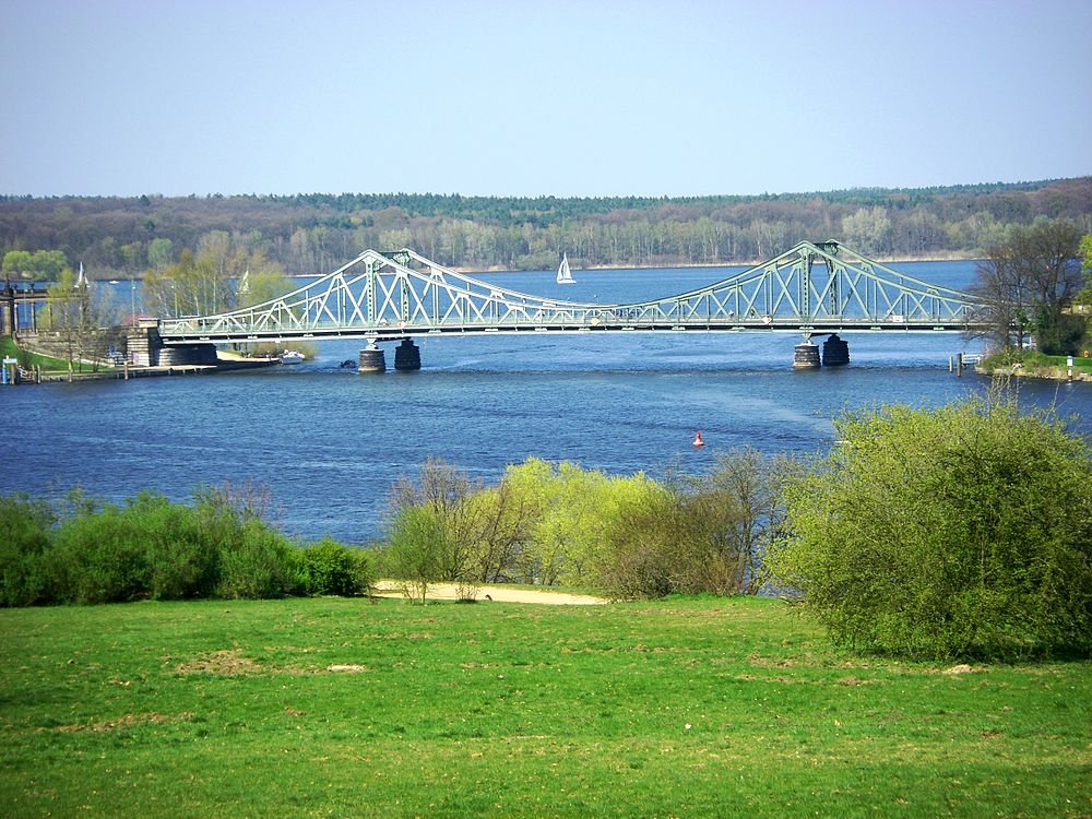 Steel truss bridge over river with greenery Scenic view of a steel truss bridge over a wide river with green grass and trees in the foreground under clear blue sky
