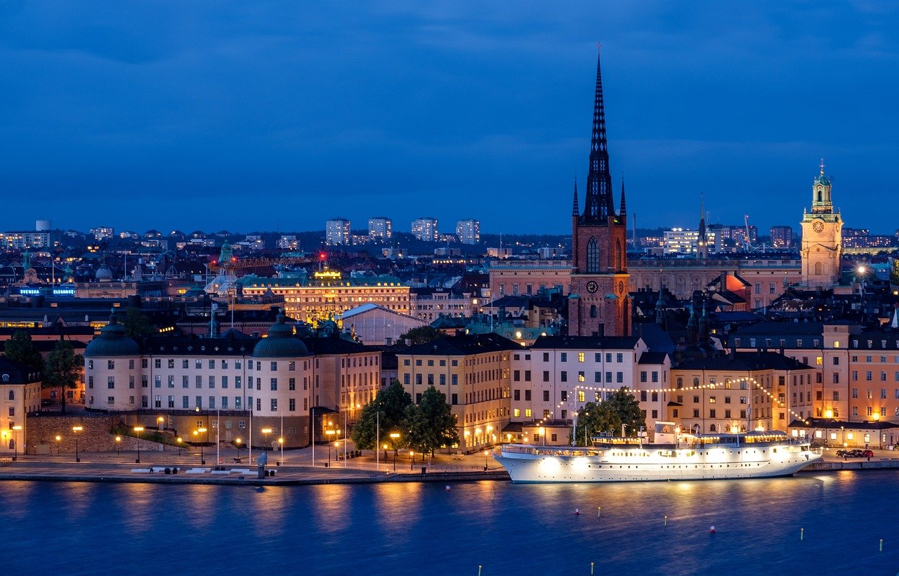 Night view of Stockholm waterfront with illuminated historic buildings, church spires, and a docked white boat reflecting on water