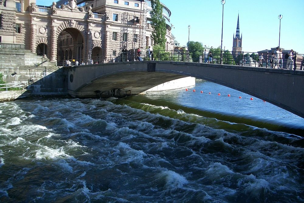 Stone arch bridge over turbulent river water with historic buildings and church spire in the background under clear sky