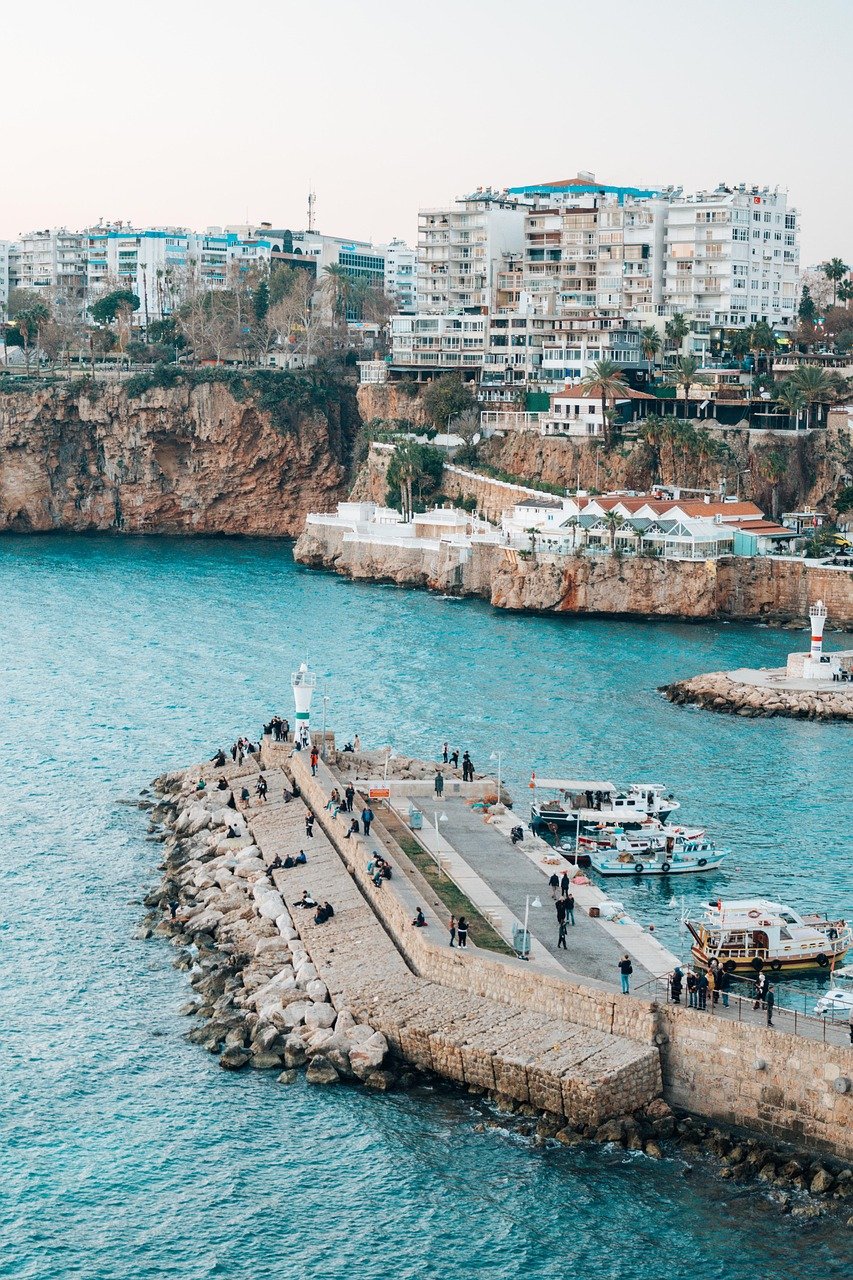 Scenic view of a stone pier with people and boats near rocky cliffs and urban buildings by the blue sea