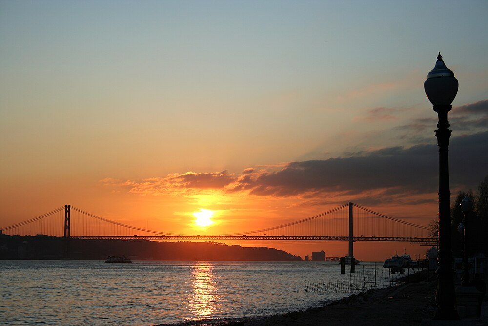 Sunset over suspension bridge and river Sunset view of a suspension bridge over a river with street lamps along the waterfront and a boat on the water