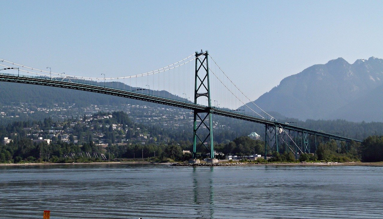 Suspension bridge over calm water with mountainous landscape and residential area in the background on a clear day