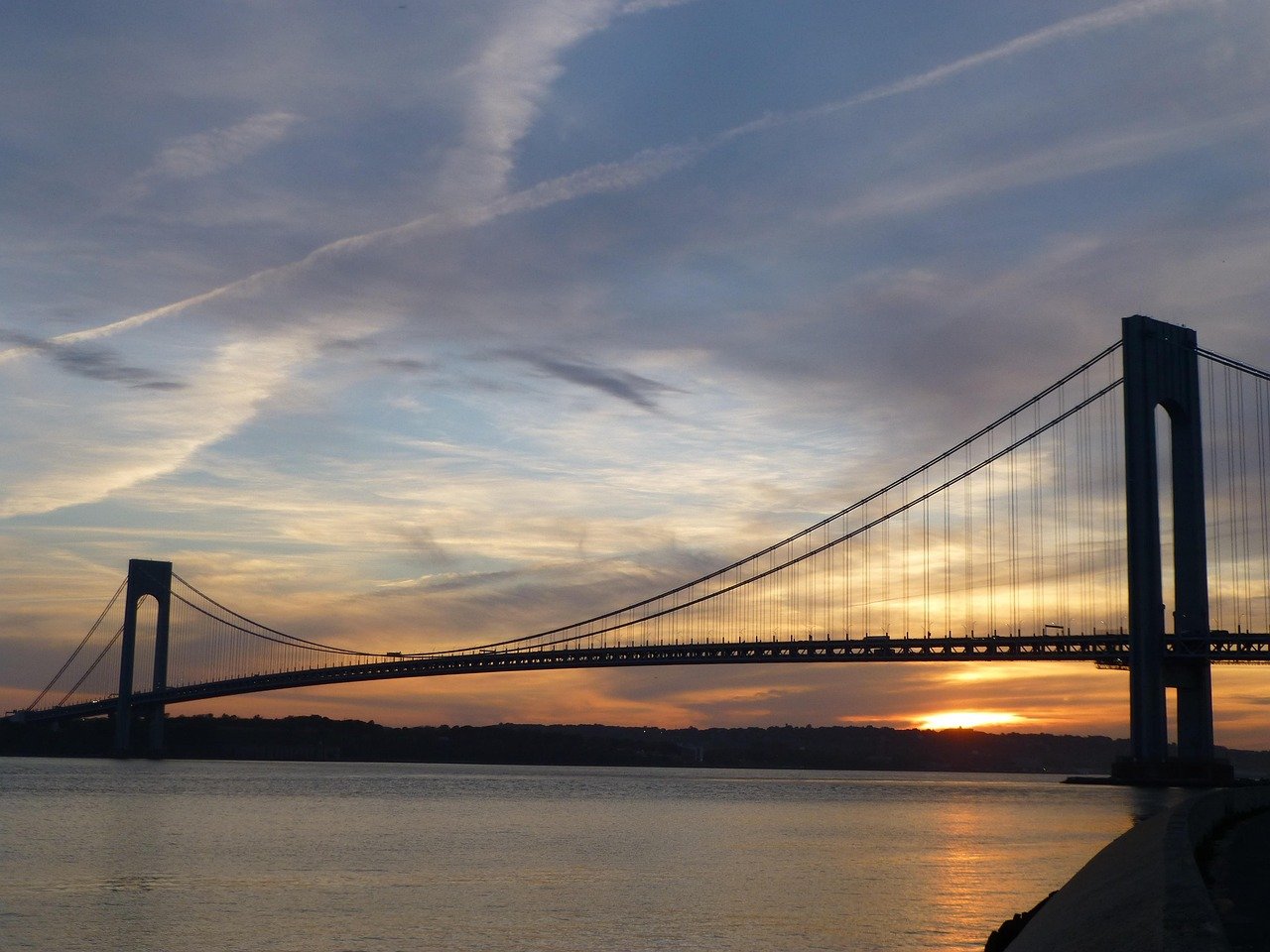 Suspension bridge at sunset over calm water with dramatic sky and silhouetted towers
