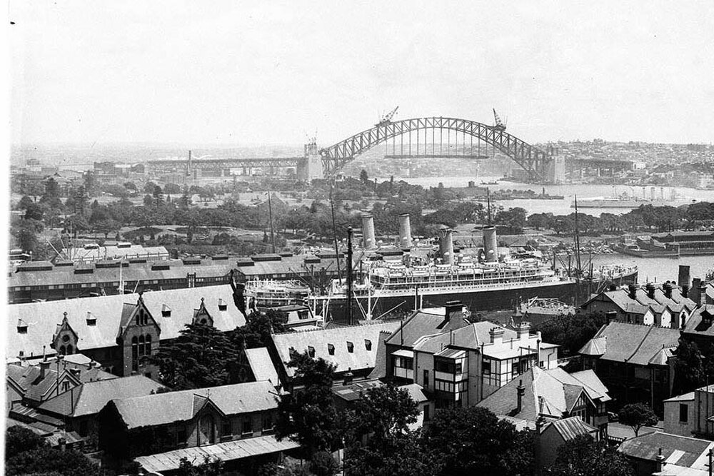 Sydney Harbour Bridge under construction with ship Historic black and white photo of Sydney Harbour with a large ship docked and Sydney Harbour Bridge under construction