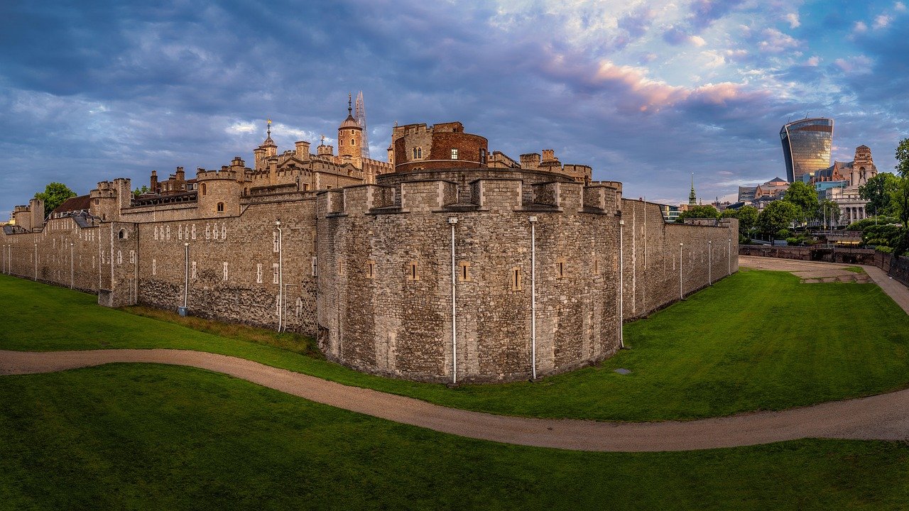 Historic Tower of London fortress with stone walls and modern city skyline under a cloudy sky at sunset