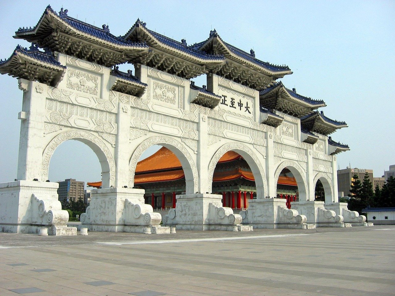 Traditional Chinese archway with intricate carvings and blue tiled roofs in a large open plaza under clear sky