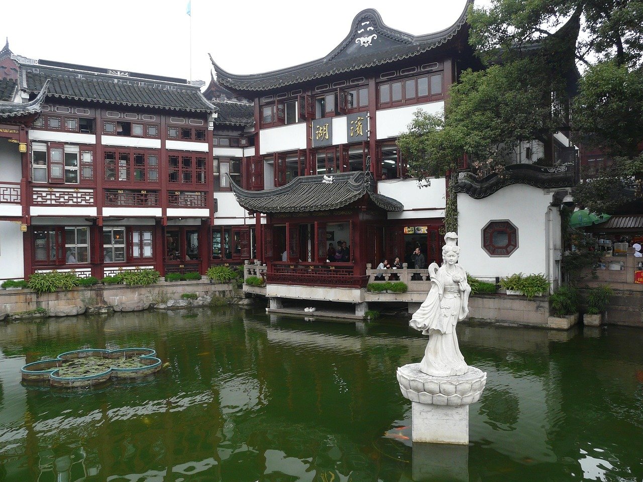 Traditional Chinese architecture with ornate roof, red wooden windows, and a white statue in a pond surrounded by greenery